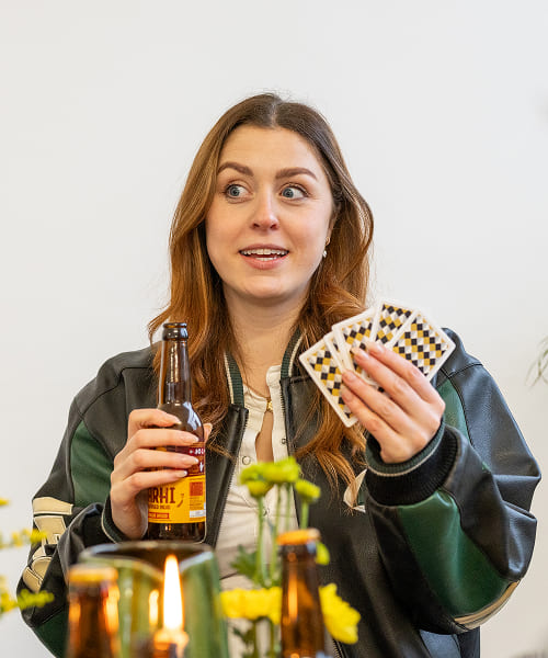 A woman holding playing cards and a bottle of Barhi date-based mead, smiling during a game night.