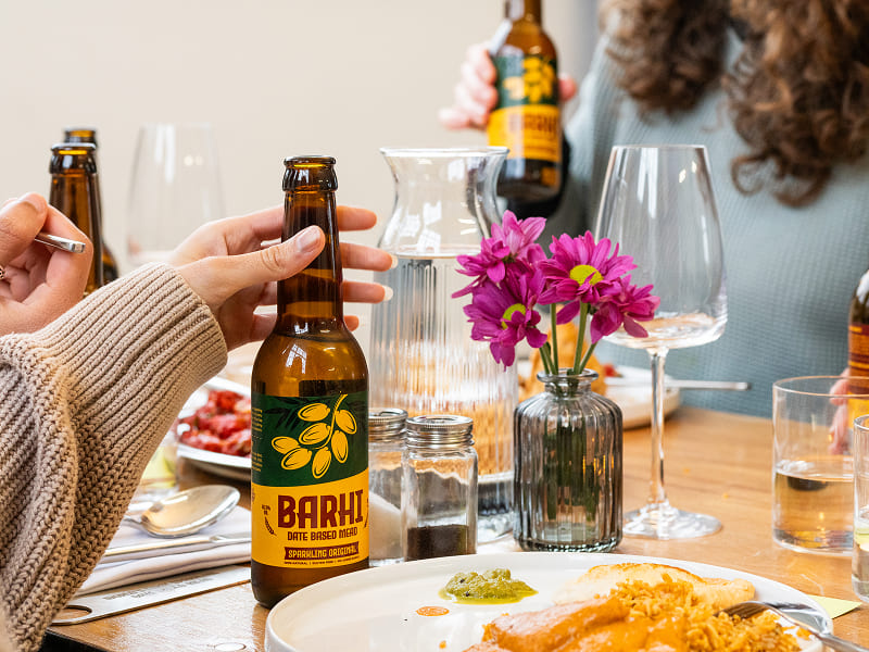 Close-up of a Barhi sparkling date-based mead bottle on a dinner table with plates, glasses, and flowers.
