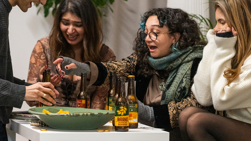 A group of friends laughing and reaching for Barhi date-based mead bottles around a table during a casual gathering.