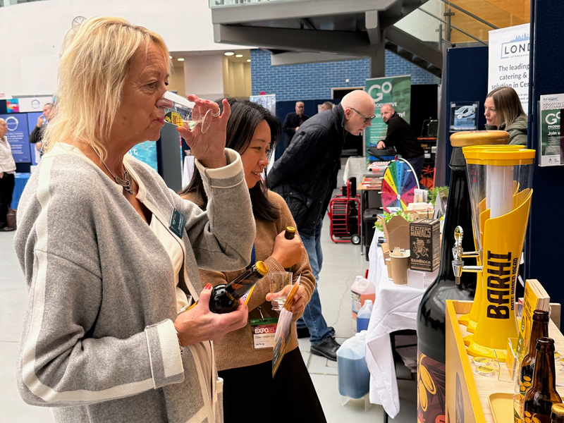 Two women tasting Barhi date-based mead at an expo booth with Barhi bottles and tap displays.