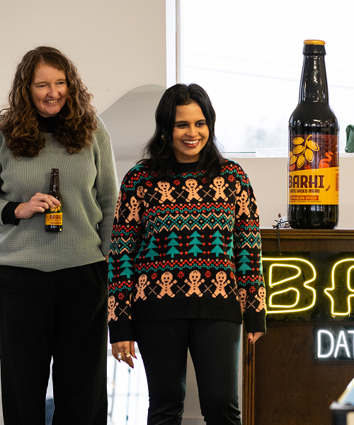 Two women standing beside a large Barhi date-based mead display bottle at an indoor event.
