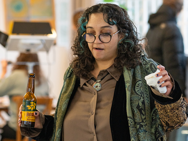 A woman holding a bottle of Barhi date-based mead at an event, looking down with a surprised expression.