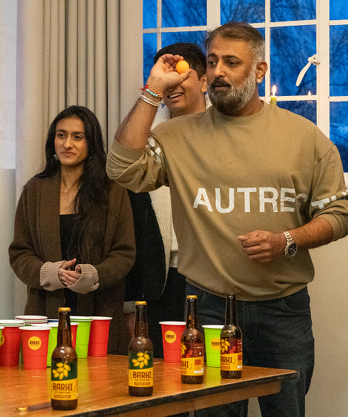 A man tossing a ping-pong ball toward cups on a table while others watch, with Barhi date-based mead bottles arranged nearby.