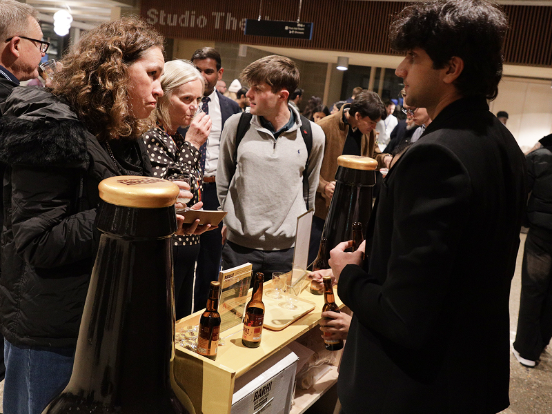 People gathered around a Barhi date-based mead tasting stand at a busy indoor event, sampling drinks and talking with a representative.