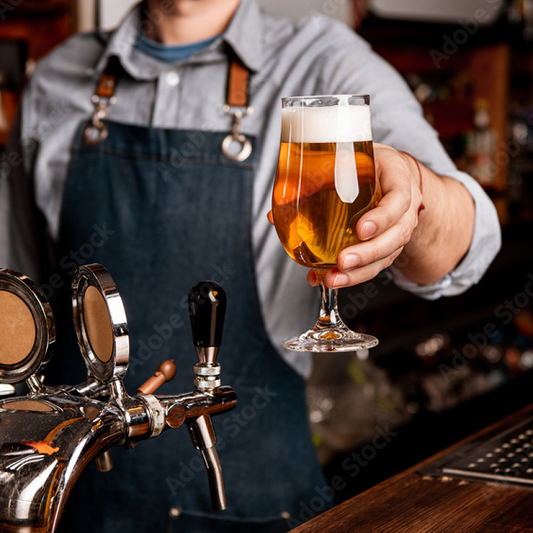 Bartender holding a freshly poured mead in a stemmed glass at a bar counter with mead taps in the foreground.