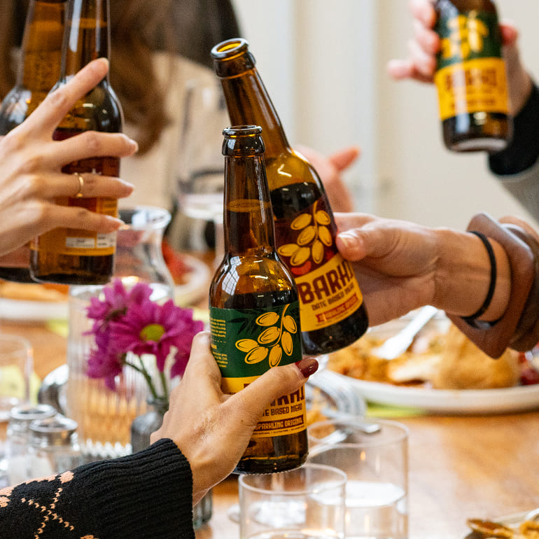 Close-up of friends clinking Barhi date-based mead bottles together over a shared meal at a dining table.