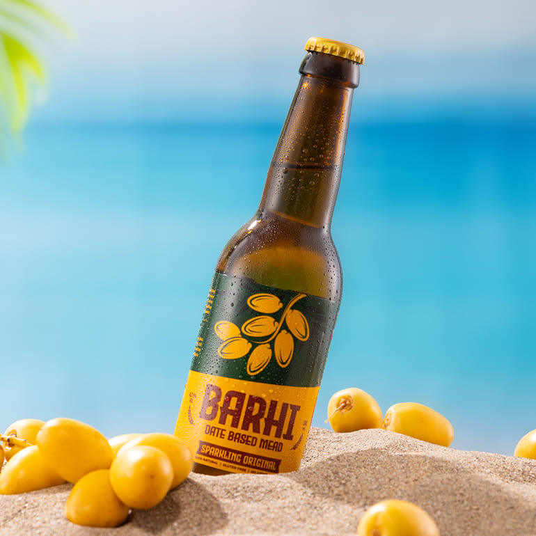 Barhi Original date-based sparkling mead bottle placed in sand with fresh yellow dates, set against a bright tropical ocean background.