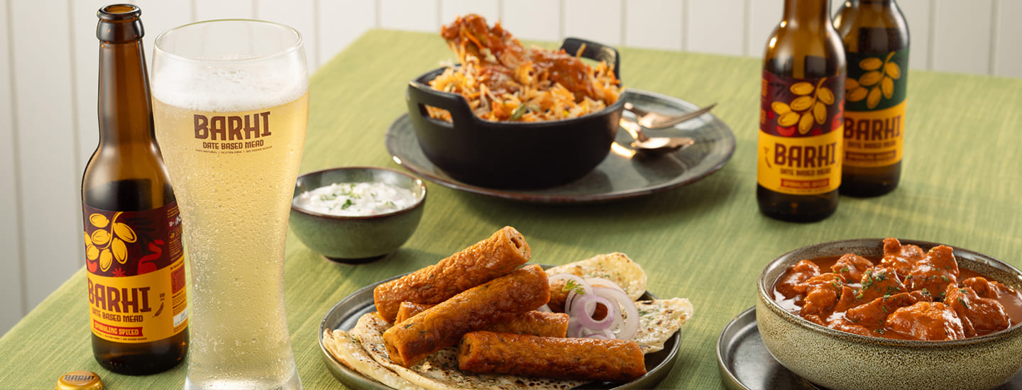 Barhi date-based sparkling mead served with Indian food dishes on a dining table, featuring a chilled glass and bottled variants.