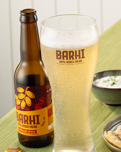 Barhi date-based sparkling mead served with Indian food dishes on a dining table, featuring a chilled glass and bottled variants.