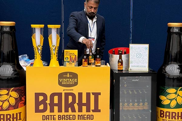 A person serving Barhi date-based mead at a promotional booth at Kingston University featuring Barhi bottles acrsoss display counters
