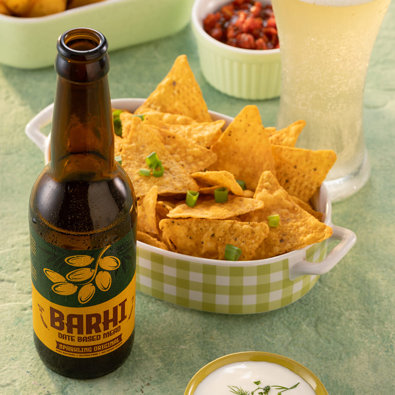 A Barhi sparkling date-based mead bottle next to a bowl of nachos and dip on a green table.