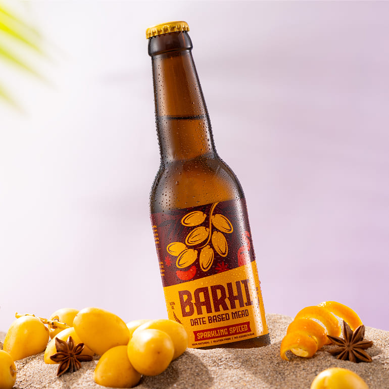 Barhi Spiced date-based sparkling mead bottle in sand, surrounded by yellow dates and whole spices, photographed against a soft pastel background.