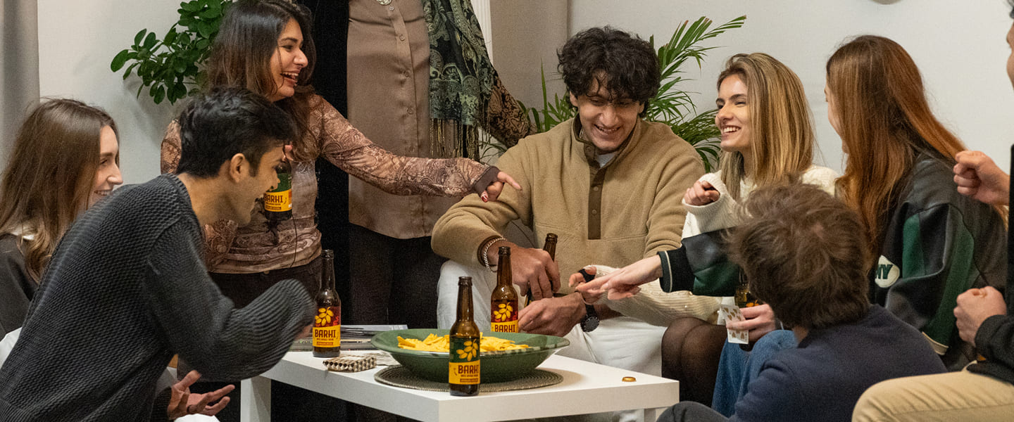 A group of friends gathered around a table indoors, laughing and playing a game while sharing Barhi date-based mead bottles and snacks