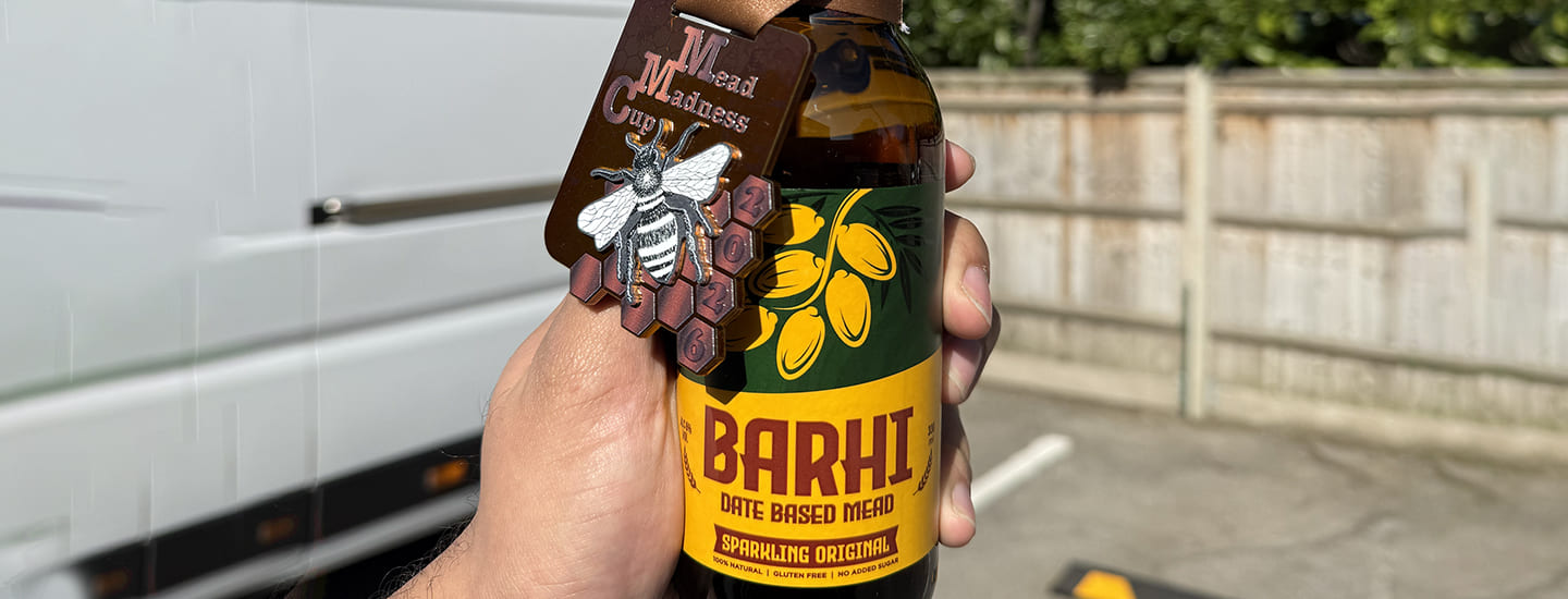 Hand holding a bottle of Barhi date-based sparkling mead with a “Mead Madness Cup” tag attached, photographed outdoors in daylight