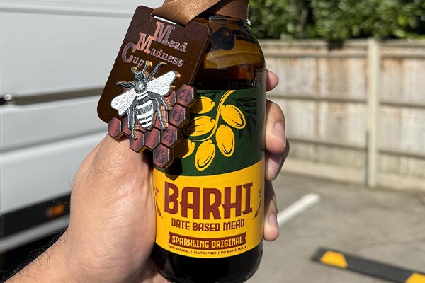 Hand holding a bottle of Barhi date-based sparkling mead with a “Mead Madness Cup” tag attached, photographed outdoors in daylight