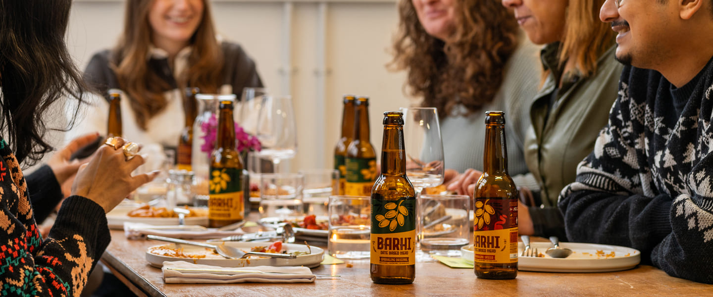 Friends seated around a dining table sharing food and bottles of Barhi date-based mead during a meal.