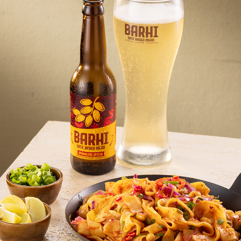 A bottle of Barhi sparkling spiced date-based mead next to a tall glass of the drink, served with a plate of spicy noodles and garnishes.