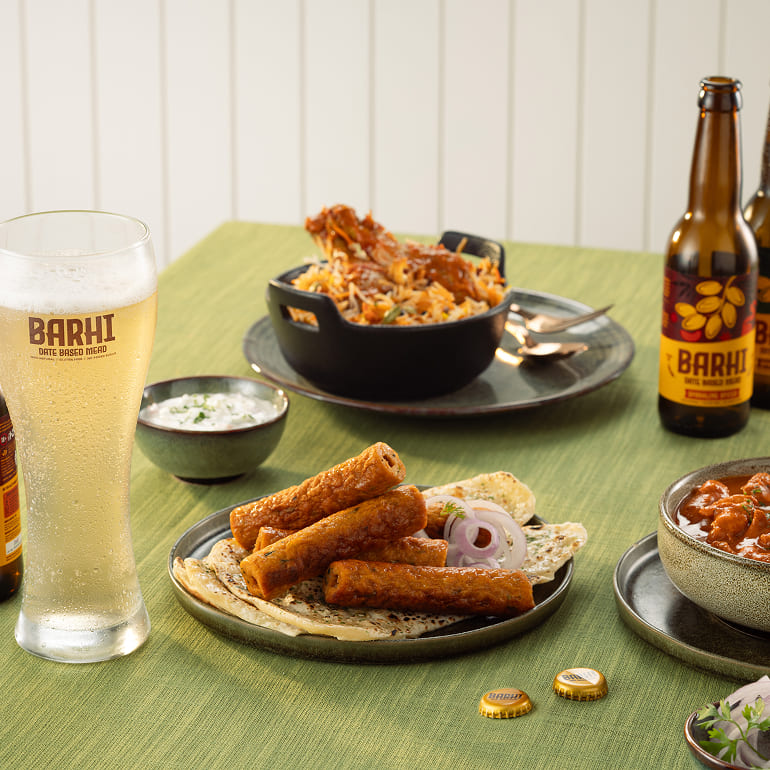 A table set with Indian-style dishes and snacks, accompanied by bottles and a glass of Barhi sparkling spiced date-based mead.