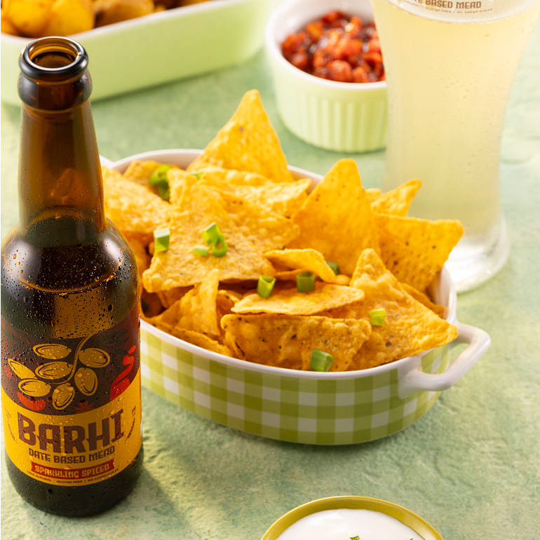 A Barhi sparkling spiced date-based mead bottle beside a bowl of tortilla chips on a green table.