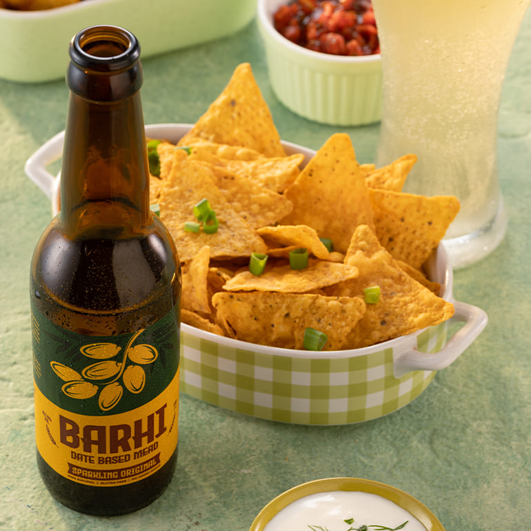 Barhi sparkling date-based mead bottle beside a bowl of tortilla chips on a table, with a glass of the drink in the background.