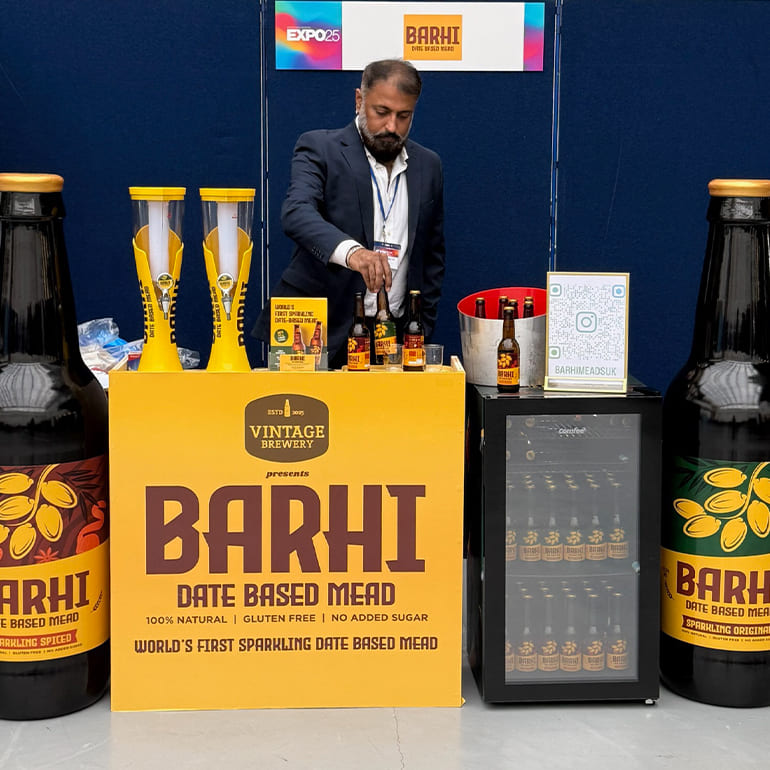 A man standing behind a Barhi date-based mead booth at an expo, with Barhi bottles, branded signage, and a display fridge.