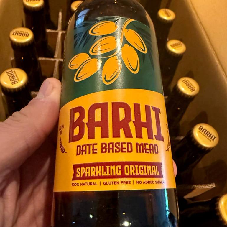 Close-up of a hand holding a Barhi sparkling date-based mead bottle, with more Barhi bottles stacked in the background.