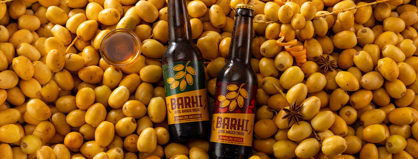 Two bottles of Barhi date-based mead resting on a bed of fresh yellow Barhi dates with spices and a small glass of mead