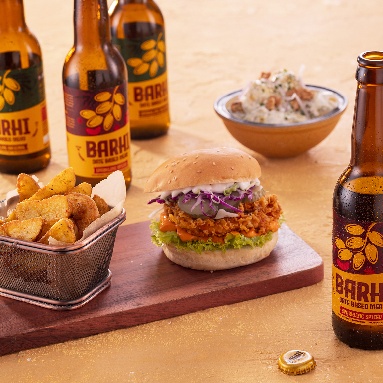 Barhi sparkling date-based mead bottles served alongside a crispy fried burger, seasoned fries, and dipping sauce on a wooden board