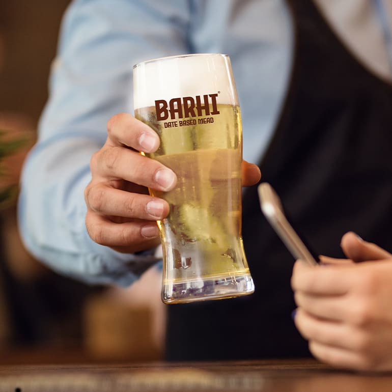 Bartender holding a freshly poured mead in a stemmed glass at a bar counter with mead taps in the foreground.