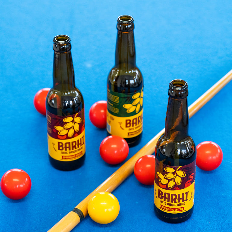 Barhi Original and Spiced date-based mead bottles arranged on a pool table with billiard balls.
