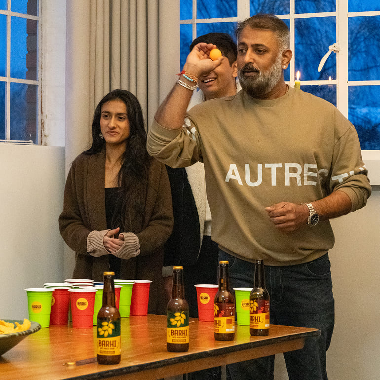 A group of people standing around a table indoors while a man playing beer pong, with several Barhi beverage bottles and colourful cups arranged on the table.