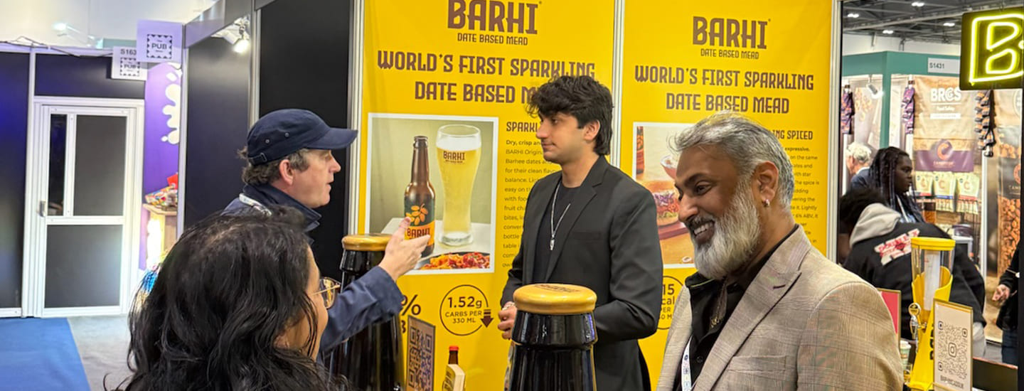 Visitors interacting at the Barhi date-based mead exhibition booth during a trade event, with branded product displays and promotional banners in the background.