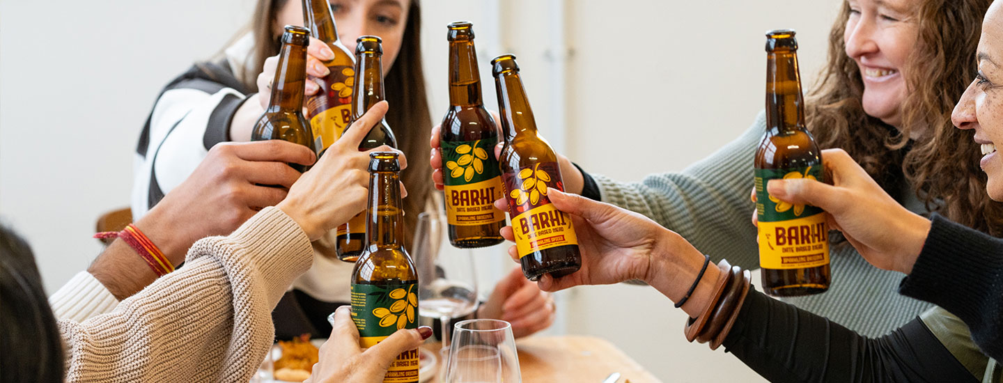 A group of people gather around a table, raising bottles of BARHI date-based mead in a celebratory toast. The bottles feature different BARHI variants - Original and Spiced