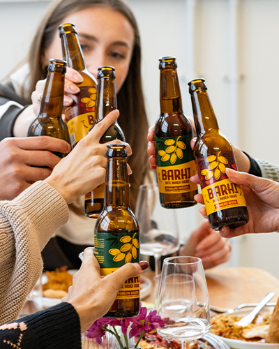 A group of people gather around a table, raising bottles of BARHI date-based mead in a celebratory toast. The bottles feature different BARHI variants - Original and Spiced