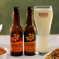 Two BARHI beverage bottles placed beside a tall branded glass filled with a light golden drink, styled with small bowls of snacks on a table against a muted green background
